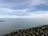 A calm seascape with an expansive body of water extending to the horizon under a partly cloudy sky. A line of distant wind turbines is visible on the horizon. In the foreground, a rocky shoreline is present with some visible greenery.