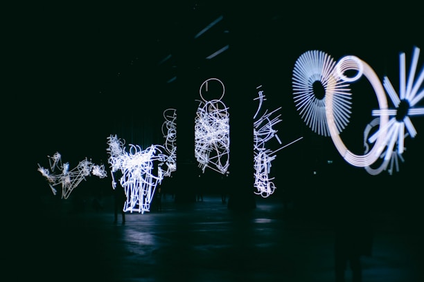 An indoor art installation with bright white light forms arranged in intricate, abstract shapes against a dark background. The designs include circular and linear patterns, creating a futuristic and dynamic atmosphere.