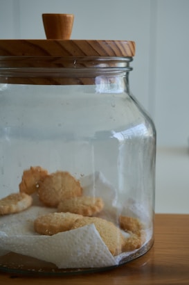 A transparent glass jar with a wooden lid contains several buttery cookies resting on a piece of white paper towel. The jar is placed on a wooden surface and the background is softly blurred.
