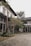 A modern architectural space with a two-story building featuring large glass windows and a flat roof. The exterior is surrounded by neatly arranged plants and trees, with a paved pathway leading up to the entrance. The overall setting is clean and minimalistic, complemented by natural greenery.