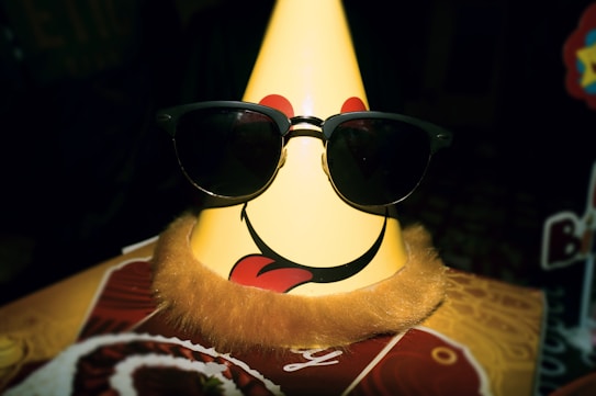 A party hat with a smiley face and tongue sticking out is accessorized with a pair of sunglasses. The hat has fluffy fur trim at the bottom. The background is dark with some indistinct objects and decorations.