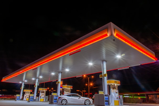 A modern gas station at night with bright overhead lights illuminating the area. The station features multiple fuel pumps under a large canopy, which is accented with red and yellow lighting. A silver car is parked next to one of the pumps. The dark night sky contrasts with the vibrant colors of the canopy and lights.