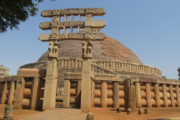 An ancient stone structure features a large dome with intricately carved gates. The architecture showcases detailed sculptural elements, including a series of columned railings and ornate decorations on the torana or gateway.