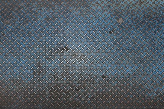 A textured metal surface featuring a repetitive diamond plate pattern with raised, angular designs. The metallic appearance is combined with a gradient of rust and dark blue tones, indicating wear and age.