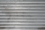 A metal roll-up shutter with horizontal slats displaying signs of rust and wear. The surface is predominantly silver with some darker smudges and spots across the slats.