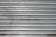 A metal roll-up shutter with horizontal slats displaying signs of rust and wear. The surface is predominantly silver with some darker smudges and spots across the slats.