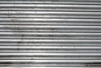 A metal roll-up shutter with horizontal slats displaying signs of rust and wear. The surface is predominantly silver with some darker smudges and spots across the slats.
