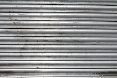 A metal roll-up shutter with horizontal slats displaying signs of rust and wear. The surface is predominantly silver with some darker smudges and spots across the slats.