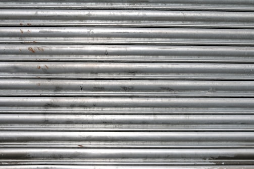 A metal roll-up shutter with horizontal slats displaying signs of rust and wear. The surface is predominantly silver with some darker smudges and spots across the slats.