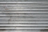 A metal roll-up shutter with horizontal slats displaying signs of rust and wear. The surface is predominantly silver with some darker smudges and spots across the slats.