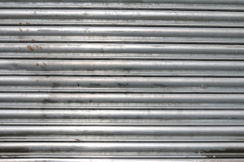 A metal roll-up shutter with horizontal slats displaying signs of rust and wear. The surface is predominantly silver with some darker smudges and spots across the slats.