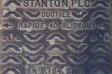 A close-up of a metal manhole cover with various raised patterns and inscriptions. The cover includes the text 'STANTON PLC', 'DUCTILE', 'RAPIDE 40 SLIDEOUT', as well as some alphanumeric codes such as 'HC036' and 'EN124 B125'. The design features numerous chevron shapes and arrows.