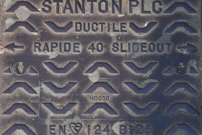 A close-up of a metal manhole cover with various raised patterns and inscriptions. The cover includes the text 'STANTON PLC', 'DUCTILE', 'RAPIDE 40 SLIDEOUT', as well as some alphanumeric codes such as 'HC036' and 'EN124 B125'. The design features numerous chevron shapes and arrows.