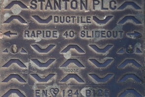 A close-up of a metal manhole cover with various raised patterns and inscriptions. The cover includes the text 'STANTON PLC', 'DUCTILE', 'RAPIDE 40 SLIDEOUT', as well as some alphanumeric codes such as 'HC036' and 'EN124 B125'. The design features numerous chevron shapes and arrows.