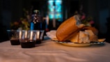 A table is set with a loaf of bread and slices on a plate, next to several plastic cups filled with red liquid. In the background, there is a dark setting with stained glass windows visible, suggesting a church or religious setting.