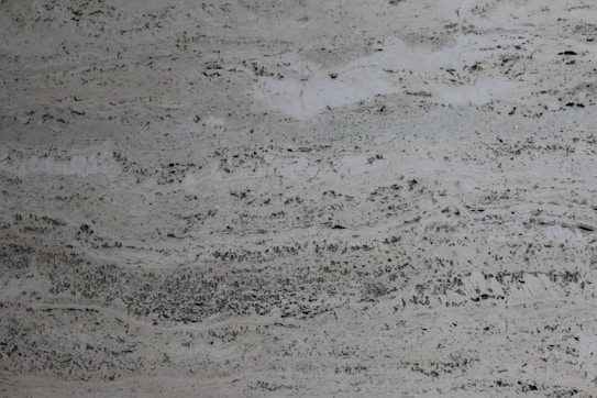 A textured stone surface with a mix of light and dark gray tones, featuring irregular patterns and natural striations creating a marble-like appearance.