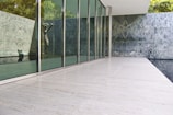 A modern architectural space featuring a sleek glass wall that reflects a stone statue inside. The area is defined by clean lines and minimalist design, with large stone tiles on the floor and wall cladding with intricate patterns on the marble surface.