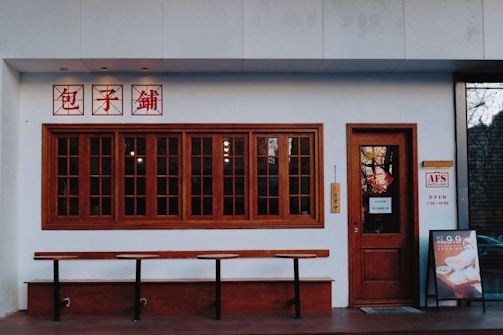 A storefront with a series of wooden-framed windows and a door, featuring signage in red characters above. There is a long wooden bench in front, and a menu or promotional sign is displayed to the right. The establishment appears to be a cafe, with a door sign indicating its opening hours.