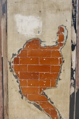 A section of an old wall with peeling plaster revealing a layer of red bricks underneath. The plaster is worn and discolored, surrounded by frayed edges and chipped areas.