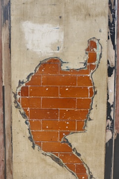 A section of an old wall with peeling plaster revealing a layer of red bricks underneath. The plaster is worn and discolored, surrounded by frayed edges and chipped areas.