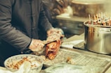 A person wearing a dark coat and gloves carves a roasted duck. The duck lies on a metal tray alongside a bowl filled with more pieces of duck. On the right side, a large pot with skewers sticking out stands on another part of the counter.