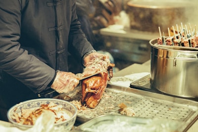 A person wearing a dark coat and gloves carves a roasted duck. The duck lies on a metal tray alongside a bowl filled with more pieces of duck. On the right side, a large pot with skewers sticking out stands on another part of the counter.