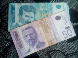 Two banknotes from the National Bank of Serbia are placed side by side. The top banknote is of 100 dinara, predominantly blue in color, and features the image of a man with a mustache. The lower banknote is of 50 dinara, primarily purple and displays another man with a beard. Both banknotes have distinctive security features and Serbian text.