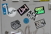 A collection of colorful stickers on a light gray surface, including a blue abstract face with eyes on one sticker, and various text-based designs featuring words like 'UGLYHOUSE', 'GREY JAM', and 'JOBZY'. Some stickers display bold typography, while others are artistic or illustrative.