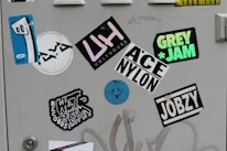 A collection of colorful stickers on a light gray surface, including a blue abstract face with eyes on one sticker, and various text-based designs featuring words like 'UGLYHOUSE', 'GREY JAM', and 'JOBZY'. Some stickers display bold typography, while others are artistic or illustrative.