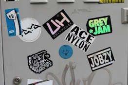 A collection of colorful stickers on a light gray surface, including a blue abstract face with eyes on one sticker, and various text-based designs featuring words like 'UGLYHOUSE', 'GREY JAM', and 'JOBZY'. Some stickers display bold typography, while others are artistic or illustrative.