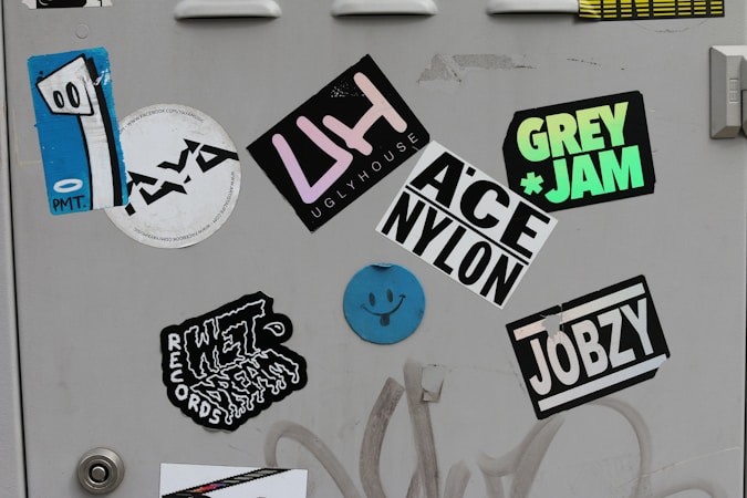 A collection of colorful stickers on a light gray surface, including a blue abstract face with eyes on one sticker, and various text-based designs featuring words like 'UGLYHOUSE', 'GREY JAM', and 'JOBZY'. Some stickers display bold typography, while others are artistic or illustrative.