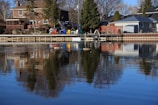 Residential houses are positioned along a calm waterfront, with a wooden deck extending over the water. Several colorful chairs and tables are arranged on the deck. Tall trees are present, and a reflection of the scene can be seen in the water.