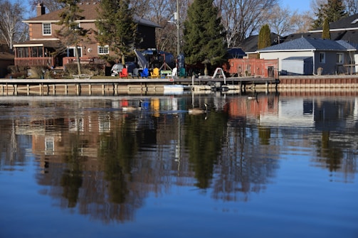 Residential houses are positioned along a calm waterfront, with a wooden deck extending over the water. Several colorful chairs and tables are arranged on the deck. Tall trees are present, and a reflection of the scene can be seen in the water.