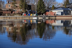 Residential houses are positioned along a calm waterfront, with a wooden deck extending over the water. Several colorful chairs and tables are arranged on the deck. Tall trees are present, and a reflection of the scene can be seen in the water.