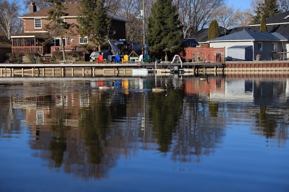 Residential houses are positioned along a calm waterfront, with a wooden deck extending over the water. Several colorful chairs and tables are arranged on the deck. Tall trees are present, and a reflection of the scene can be seen in the water.