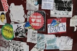 A collection of peeling and overlapping stickers on a surface, featuring a variety of designs and texts. Some stickers are worn out, and the phrases include humorous or rebellious messages. The surface appears weathered and adds to the chaotic arrangement.