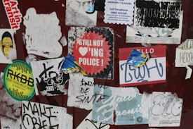 A collection of peeling and overlapping stickers on a surface, featuring a variety of designs and texts. Some stickers are worn out, and the phrases include humorous or rebellious messages. The surface appears weathered and adds to the chaotic arrangement.