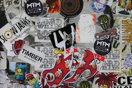 A vibrant collage of various stickers and posters layered on a surface, featuring diverse typography, logos, graphic illustrations, and cartoon characters. Some prominent logos include 'Slow Jams,' 'Timber Boards,' and 'Twizted Movements.' There are illustrations of a sinister cartoon figure and other abstract designs.