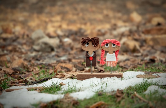 Two handmade dolls with large eyes and yarn hair are positioned on a small wooden base. The doll on the left has brown hair and is dressed in a dark outfit, while the one on the right has pink hair and wears a white dress with pink stripes. They are set against a background that features green grass and patches of snow with dry leaves scattered around.