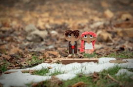 Two handmade dolls with large eyes and yarn hair are positioned on a small wooden base. The doll on the left has brown hair and is dressed in a dark outfit, while the one on the right has pink hair and wears a white dress with pink stripes. They are set against a background that features green grass and patches of snow with dry leaves scattered around.