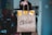 A person is holding a brown paper McCafé bag with takeaway food tags. The background indicates an indoor setting with blurred objects and decor, hinting at a casual, home-like atmosphere.