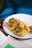 A hot dog topped with sliced jalapenos, pickled carrots, and drizzles of mustard and perhaps a spicy sauce. It is placed on a white plate with a blue rim, set on a wooden surface. There's a small bowl with what looks like a dipping sauce next to the plate.
