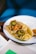 A hot dog topped with sliced jalapenos, pickled carrots, and drizzles of mustard and perhaps a spicy sauce. It is placed on a white plate with a blue rim, set on a wooden surface. There's a small bowl with what looks like a dipping sauce next to the plate.