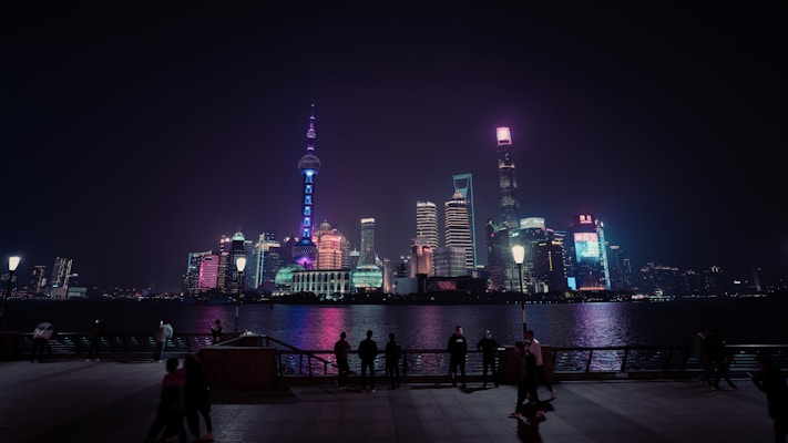 A nighttime cityscape with a prominent skyline filled with illuminated skyscrapers. Bright neon lights reflect off the water in the foreground, creating a vibrant atmosphere. Silhouettes of people are visible along the waterfront, adding a sense of scale and activity.