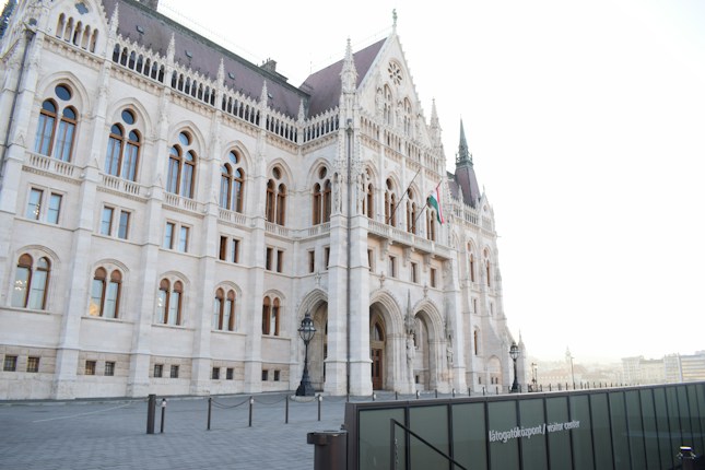 A grand, gothic-style building with intricate architectural details, featuring pointed arches, tall windows, and ornate spires. The structure is made of light stone, with a prominent Hungarian flag displayed near its entrance. The foreground has a paved area lined with bollards and a sign indicating a visitor center.