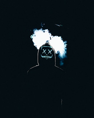 A person in a dark hoodie with a glowing neon mask featuring stitched mouth and X&rsquo;s for eyes. Smoke or vapor billows around the head, creating an eerie and mysterious atmosphere.