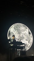 A musician is performing on stage with a keyboard setup in front of a large, black and white image of a planet, which resembles Earth. The overall lighting is dim, creating a dramatic contrast between the performer and the background.