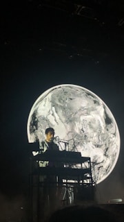 A musician is performing on stage with a keyboard setup in front of a large, black and white image of a planet, which resembles Earth. The overall lighting is dim, creating a dramatic contrast between the performer and the background.