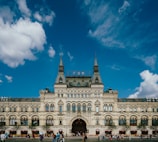 A historic, ornate building with intricate architectural details and large arched windows. The structure features multiple spires and a gabled roof, with a sign on top in Cyrillic script. People are gathered around the entrance and walking in the open square in front of the building. The sky above is clear with a few scattered clouds.