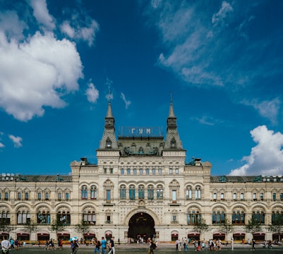 A historic, ornate building with intricate architectural details and large arched windows. The structure features multiple spires and a gabled roof, with a sign on top in Cyrillic script. People are gathered around the entrance and walking in the open square in front of the building. The sky above is clear with a few scattered clouds.